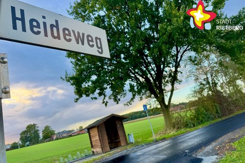 Ein Straßenschild mit der Aufschrift "Heideweg" ragt von links in das Bild, während im Hintergrund eine frisch asphaltierte Straße zu sehen ist.