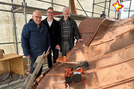 Matthias Setter, Fachbereichsleiter Bauen, Bürgermeister Andreas Sunder und Michael Kaune von der Hochbauabteilung der Stadt Rietberg stehen auf einem Gerüst auf Höhe des Dachs der Kapelle. Ein Großteil ist bereits mit Kupferplatten bestückt.