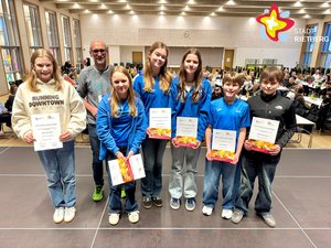 Sechs Jugendliche und ein älterer Mann stehen nebeneinander auf einer Bühne und schauen in die Kamera.