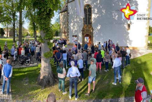 Hunderte Menschen stehen im Halbrund unter schattigen Bäumen vor einer Kirche und lauschen dem Bürgermeister.