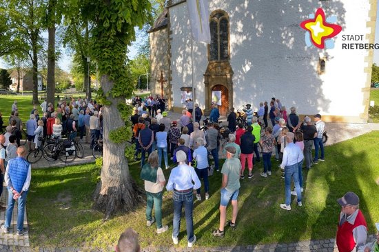 Hunderte Menschen stehen im Halbrund unter schattigen Bäumen vor einer Kirche und lauschen dem Bürgermeister.