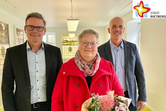 Bürgermeister Andreas Sunder und Michael Schlüter stehen links und rechts der langjährigen Mitarbeiterin Karin Schniedertöns, die zum 40-jährigen Dienstjubiläum einen Blumenstrauß erhielt, den sie in der linken Hand hält.