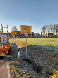 Am Straßengraben steht ein gelbes Ortseingangsschild mit der Aufschrift "Geschlossene Ortslage, Ersatz-Ortstafel, Kreis Gütersloh".