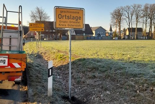Am Straßengraben steht ein gelbes Ortseingangsschild mit der Aufschrift "Geschlossene Ortslage, Ersatz-Ortstafel, Kreis Gütersloh".