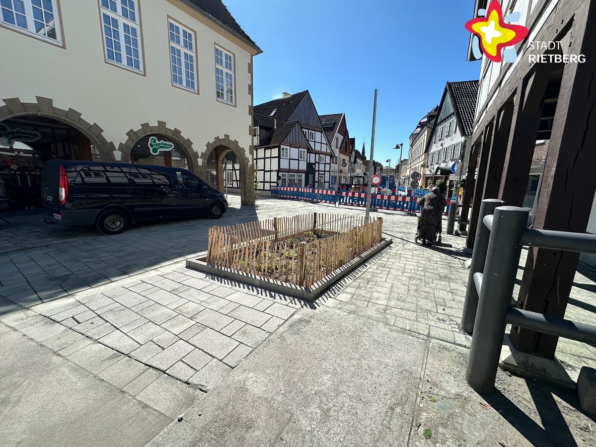 Ein großes dunkles Auto fährt im Schatten eines alten Hauses über eine frisch gepflasterte Straße. Im Vordergrund befindet sich ein niedrig eingezäuntes Blumenbeet.