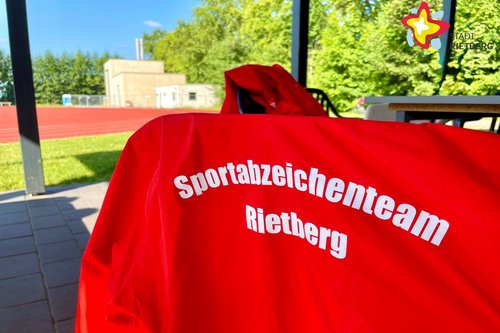 Eine rote Jacke mit der weißen Aufschrift "Sportabzeichenteam Rietberg" hängt über einem Stuhl.