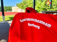 Eine rote Jacke mit der weißen Aufschrift "Sportabzeichenteam Rietberg" hängt über einem Stuhl.
