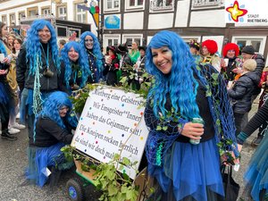 Vier Frauen mit langen blauen Haaren und Fischernetzen um die Schultern stehen um einen Bollerwagen mit der Aufschrift "Rietberg eine Geisterstadt. Wir Emsgeister*innen haben das Gejammer satt! Die Kneipen rocken herrlich, da sind wir doch mal ehrlich."