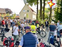 Zu sehen sind viele Radfahrer, die neben ihren Rädern stehen, weil der Bürgermeister gerade am Dorfplatz etwas erklärt.