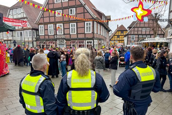Drei uniformierte Personen blicken auf eine feiernde Menge auf der Straße. Auf ihren gelben Westen lässt sich "Ordnungsamt" lesen.