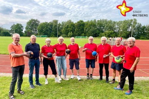 Mehrere Frauen und Männer, überwiegend in roten Shirts, stehen nebeneinander auf einem Sportplatz und halten Bälle in den Händen.