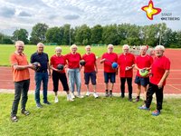 Mehrere Frauen und Männer, überwiegend in roten Shirts, stehen nebeneinander auf einem Sportplatz und halten Bälle in den Händen.