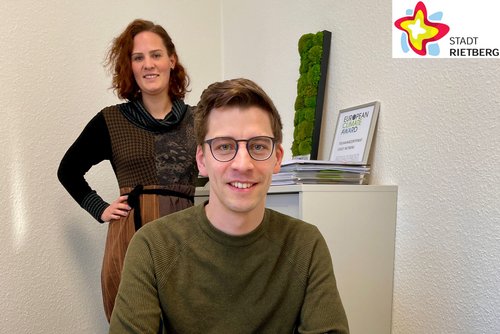 Klimaschutzmanagerin Svenja Schröder steht hinter ihrem neuen Kollegen Lukas Schmidt, der hinter seinem Schreibtisch steht.