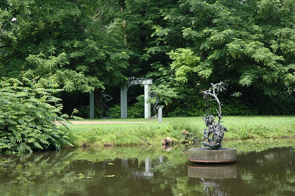 Mehrere Skulpturen stehen in einem Park, im Vordergrund ein Gewässer.