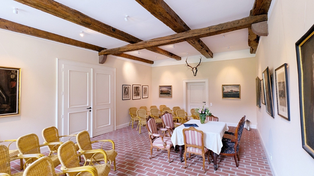 Leere Korbstühle sind in Reihen aufgestellt. Ein dunkler Tisch mit vier antiken Stühlen drumherum steht am Rand. An der Wand hängen altertümliche Bilder.