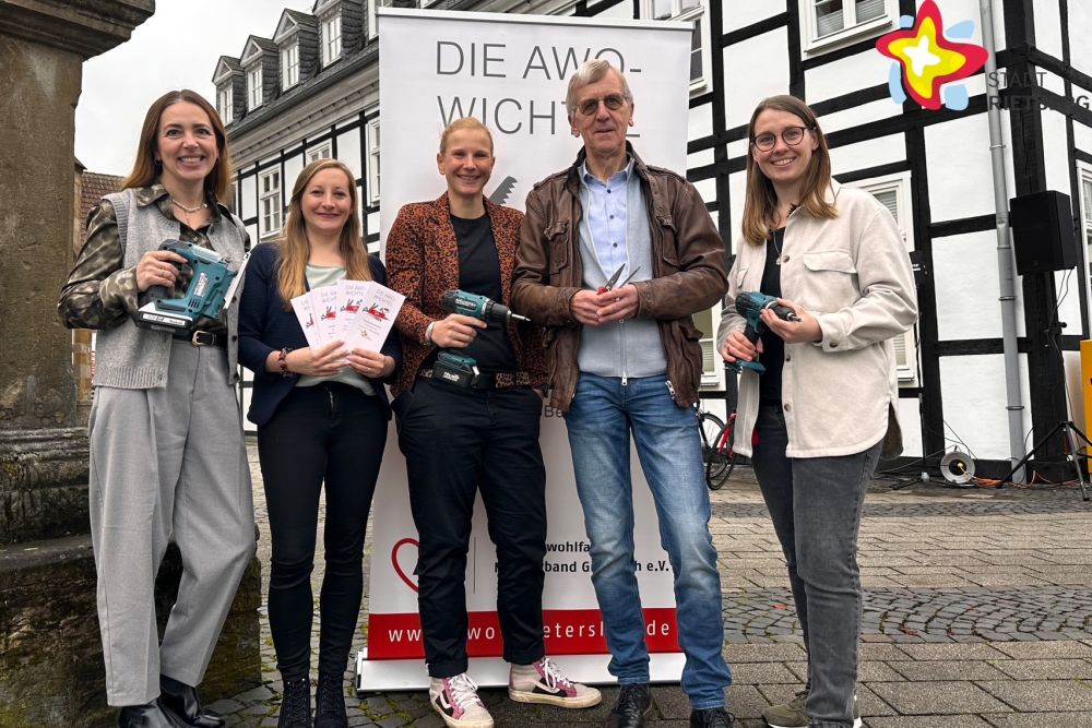 Organisatoren und ein AWO-Wichtel stehen vor einem Werbebanner mit der Aufschrift "AWO-Wichtel" und halten Werkzeug und Broschüren in der Hand.