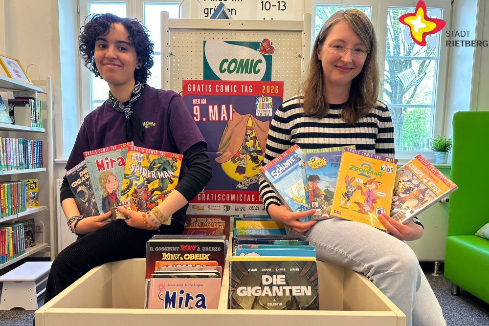 Bibliotheksleiterin Jennifer Bader (rechts) und Mitarbeiterin Laila Elgabarty sitzen in der Stadtzbibliothek rechts und links auf der Kante eines Bücherregals, das mit Comics gefüllt ist, und halten Comics wie "Spiderman", "Zoomania" oder "Mira" in der Hand.